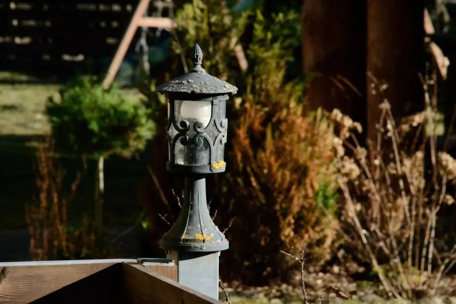 A weathered, lichen-covered decorative lantern-style post light mounted on a wooden fence post, showing years of outdoor wear in daylight.