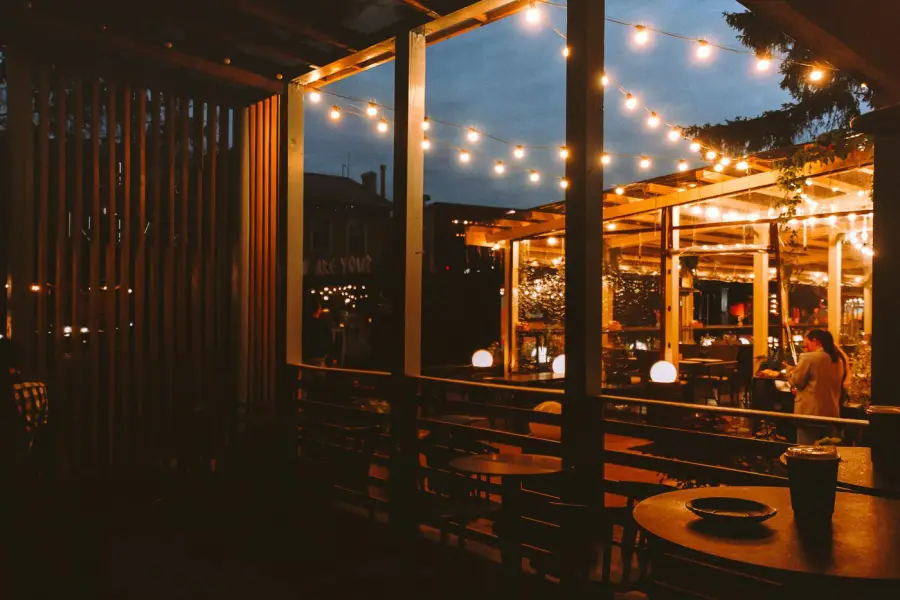 A string-light-covered outdoor patio pergola at dusk, with warm bulb lights strung overhead and globe pendant lights on café tables, diners seated in the background.
