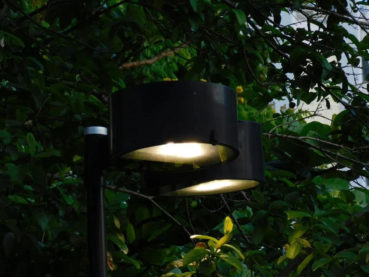 A modern black pole-mounted light fixture with dual angled lamp heads glowing warm white, mounted against dense leafy foliage at dusk with a small sensor cap visible near the top of the pole.