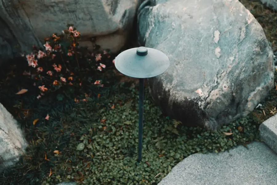 A weathered bronze mushroom-cap path light inspected in daylight, standing unlit in a rock garden groundcover bed beside large boulders and a flowering shrub.